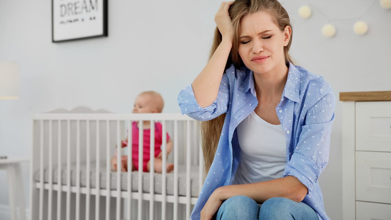 frustrated woman sitting next to a crib with infant sitting inside the crib
