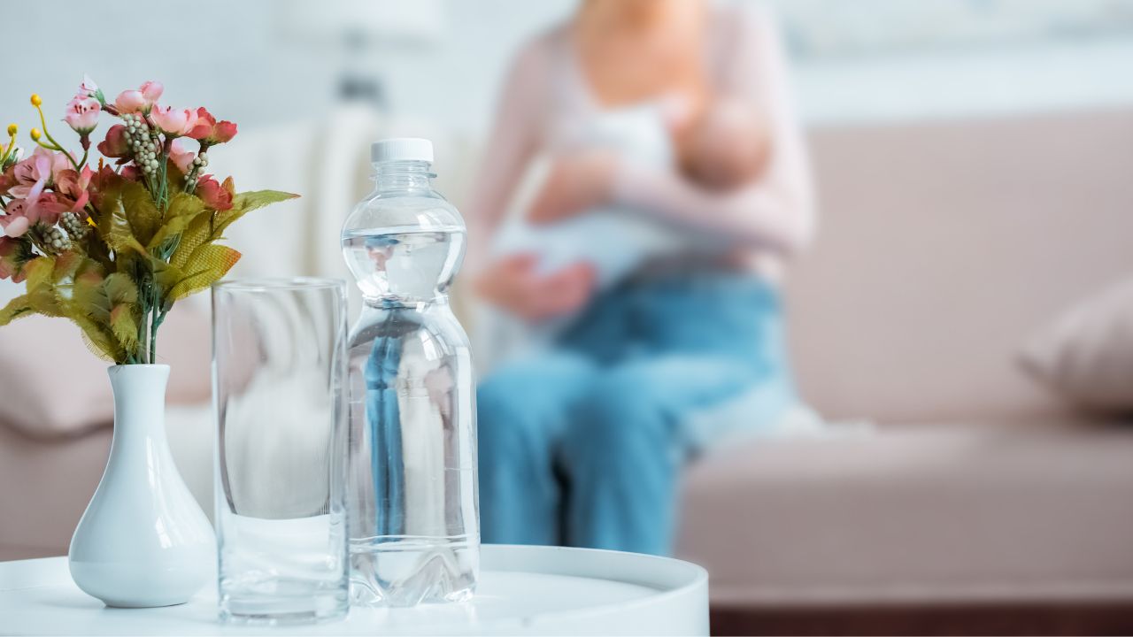 mom feeding infant in the background with a closeup of a bottle of water - one of the items on my breastfeeding station checklist